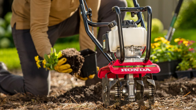 Closeup of tiller in flowerbed. In the background, a man is transplanting flowers from a pot to the flowerbed.