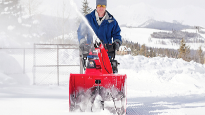 Front-facing image of man in blue jacket pushing red honda snowblower. Snow landscape in background. Trees and mountains in the distance.